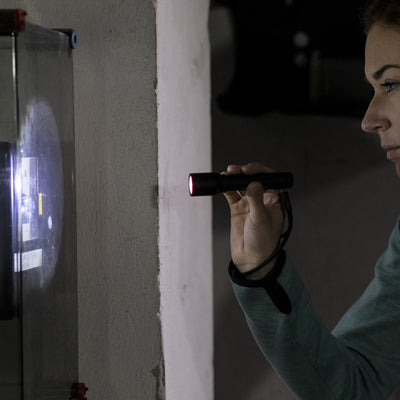 P5 Torch held by a person in a teal sleeve a small black flashlight with a red front light in a dim industrial setting