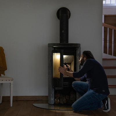 P7R Core Torch held by a man kneeling beside a wood stove in a dim living room with warm stove light