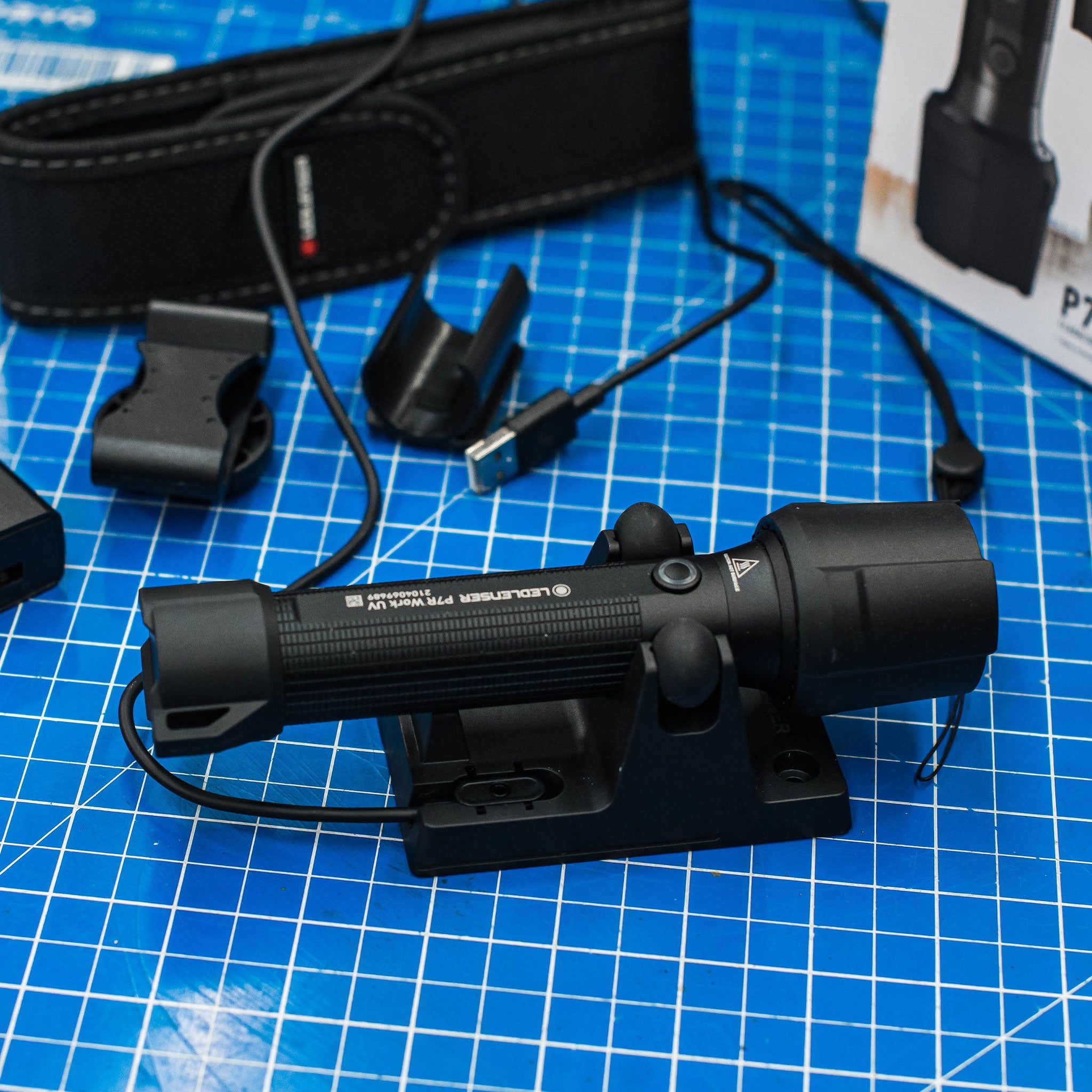 P7R Work UV Torch mounted in its charging cradle on a blue grid work surface with USB charging cable and accessories nearby