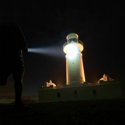 Ledlenser X21R Torch at night with a silhouetted person pointing a beam toward a lit lighthouse