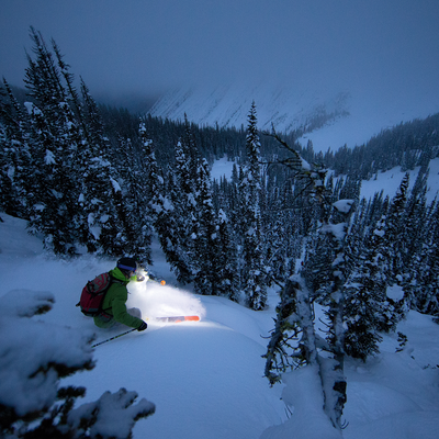 Ledlenser XEO19R Rechargeable Headlamp worn by a backcountry skier on a snowy slope with pine trees at dusk
