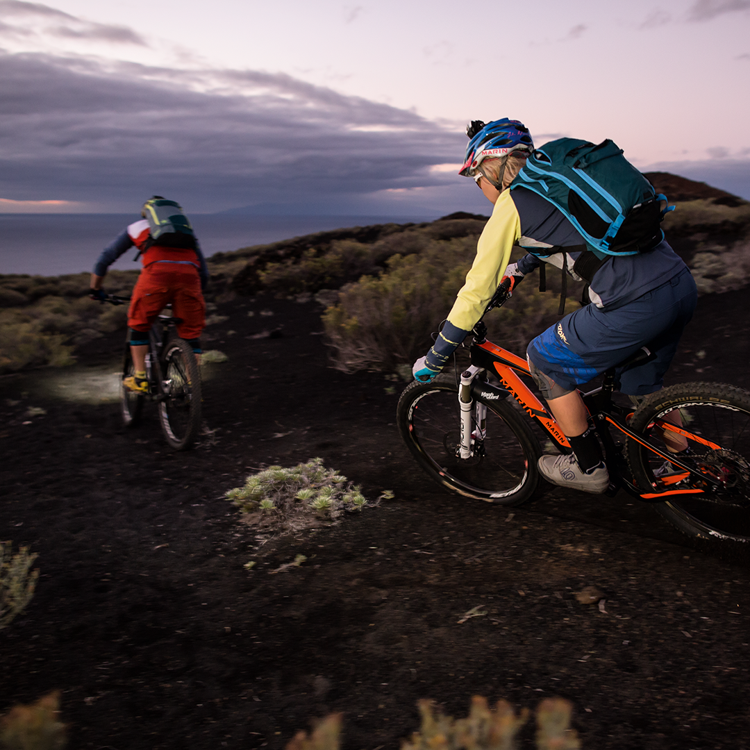 XEO19R White Rechargeable Headlamp mounted on a rider’s helmet during dusk mountain biking on a rocky trail with another rider nearby