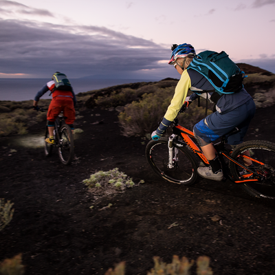 XEO19R White Rechargeable Headlamp mounted on a rider’s helmet during dusk mountain biking on a rocky trail with another rider nearby