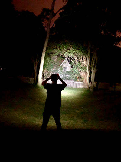 MH11 Blue Outdoor Headlamp worn on head by a person in a dark yard with the beam lighting grass and a house in the background