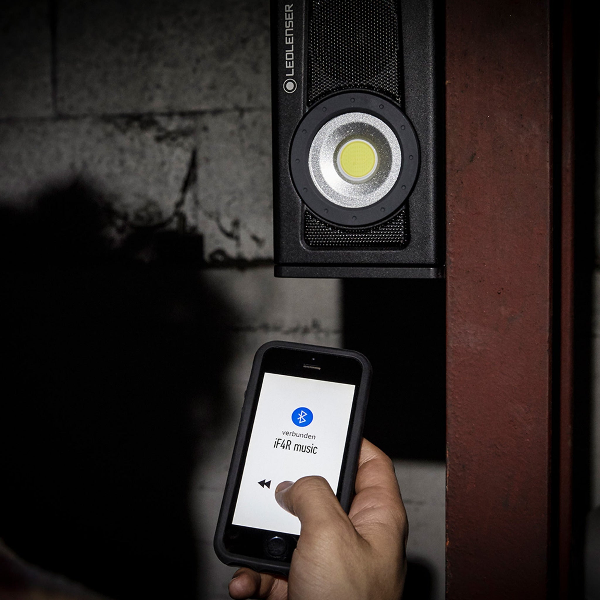 Ledlenser iF4R Music Spotlight mounted on a wall with circular LED head and a hand holding a smartphone showing Bluetooth music control