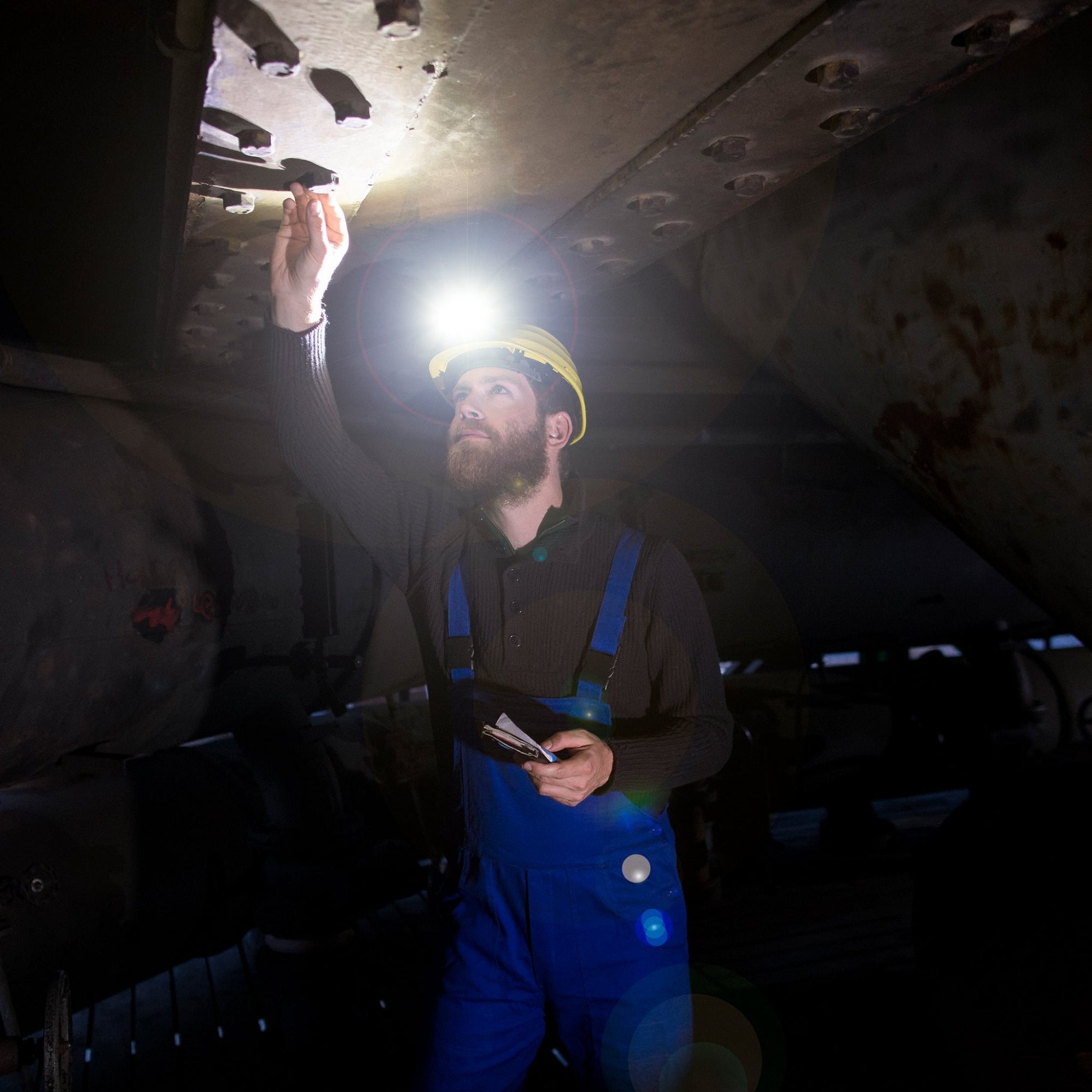 iH9R Headlamp mounted on a worker's yellow helmet illuminating an industrial ceiling while he checks bolts in blue overalls